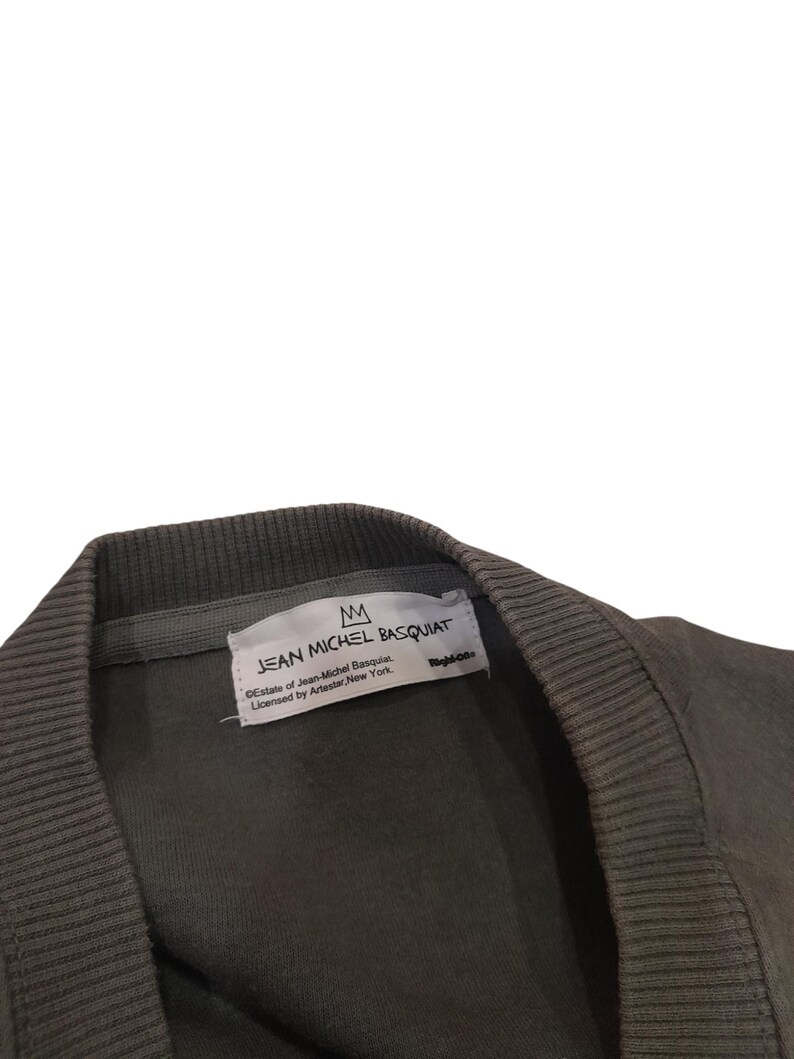 May include: Close-up of a dark olive green sweatshirt collar. A white tag displays the text "JEAN MICHEL BASQUIAT" with a crown logo. The tag also includes licensing information from Artestar, New York.
