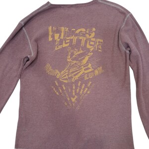 May include: A mauve long-sleeve thermal shirt with exposed white stitching. The back features a distressed yellow graphic of a skeleton riding a bomb with the text WINGS LETTER and SKULLS LUCK, along with a pattern of small falling bomb icons.