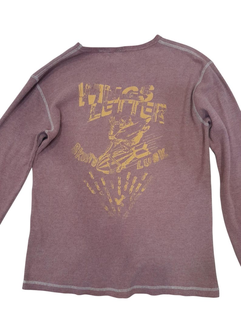 May include: A mauve long-sleeve thermal shirt with exposed white stitching. The back features a distressed yellow graphic of a skeleton riding a bomb with the text WINGS LETTER and SKULLS LUCK, along with a pattern of small falling bomb icons.