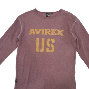 May include: A long-sleeve thermal shirt in a muted mauve color with a crew neckline. The chest features the text AVIREX US in a distressed yellow font. The garment has visible white contrast stitching along the seams and a small black brand tag.