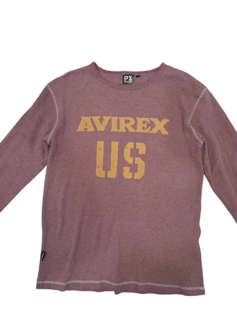 May include: A long-sleeve thermal shirt in a muted mauve color with a crew neckline. The chest features the text AVIREX US in a distressed yellow font. The garment has visible white contrast stitching along the seams and a small black brand tag.
