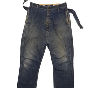 May include: High-waisted blue denim pants featuring a vertical button-fly closure, side pockets, and an integrated adjustable belt with snap buttons. The fabric has a faded wash, a small distressed area on the right leg, and a Kapital brand label.