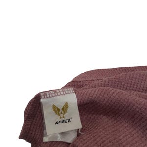 May include: A mauve-colored waffle-knit fabric garment featuring a white rectangular interior tag. The tag displays a gold-colored graphic of wings and a star above the text AVIREX in black capital letters with a registered trademark symbol.