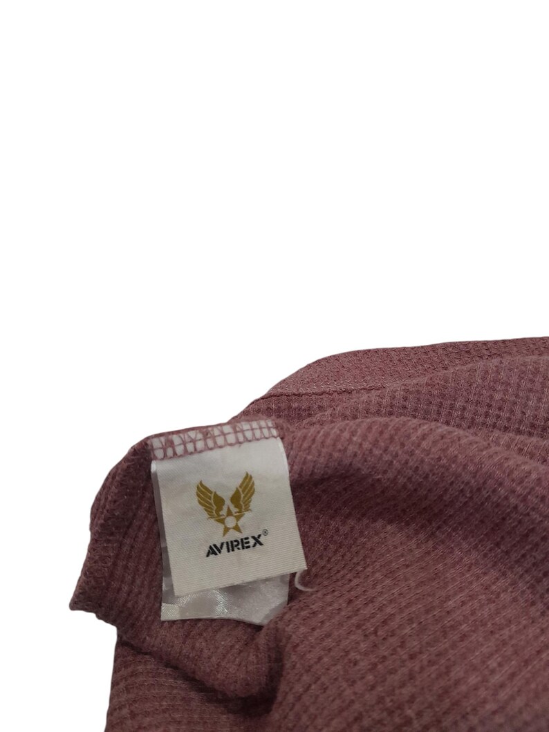 May include: A mauve-colored waffle-knit fabric garment featuring a white rectangular interior tag. The tag displays a gold-colored graphic of wings and a star above the text AVIREX in black capital letters with a registered trademark symbol.