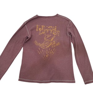 May include: A long-sleeve mauve shirt with visible white stitching along the seams. The back features a gold-toned graphic print with the text "WINGS LETTER" above an illustration of a winged object and the word "LUCK" below the graphic.