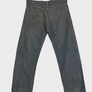May include: Faded blue denim jeans with classic five-pocket styling. The back pockets feature a distinctive curved stitch design. The jeans are slightly worn, showing signs of use. The waistline has a red accent.