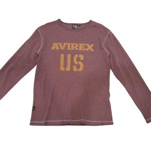 May include: A long-sleeve maroon shirt with a crew neckline and visible white stitching along the seams. The chest features the text AVIREX and US in a distressed tan font. A small black brand tag is attached to the lower right side hem.