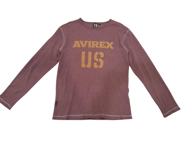 May include: A long-sleeve maroon shirt with a crew neckline and visible white stitching along the seams. The chest features the text AVIREX and US in a distressed tan font. A small black brand tag is attached to the lower right side hem.
