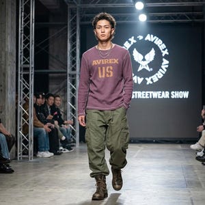 May include: A long-sleeve purple thermal shirt with the text AVIREX US printed in gold on the chest, paired with olive green cargo pants featuring side pockets and brown leather lace-up boots, worn during a fashion show with a branded backdrop.