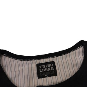 Puede incluir: Una prenda negra con cuello redondo y forro interior a rayas finas blancas y negras. Una etiqueta de tela rectangular negra est&aacute; cosida en el centro del cuello, mostrando el texto blanco Y'S FOR LIVING BASICS en una fuente sin remates.