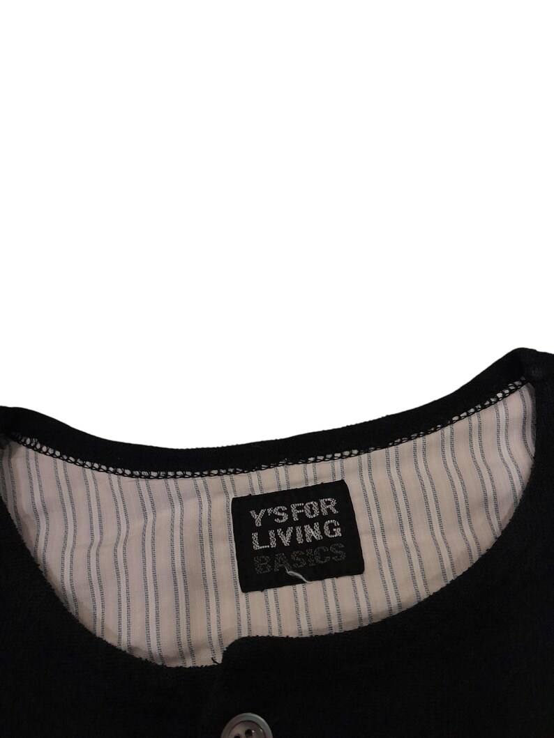 Puede incluir: Una prenda negra con cuello redondo y forro interior a rayas finas blancas y negras. Una etiqueta de tela rectangular negra est&aacute; cosida en el centro del cuello, mostrando el texto blanco Y'S FOR LIVING BASICS en una fuente sin remates.