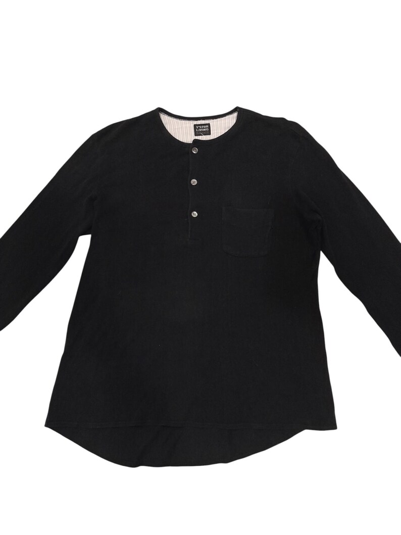 Puede incluir: Una camiseta henley negra de manga larga con una tapeta de tres botones, un bolsillo en el pecho y un dobladillo redondeado. El forro interior del cuello es blanco con finas rayas verticales negras e incluye una etiqueta que dice Y's FOR LIVING.