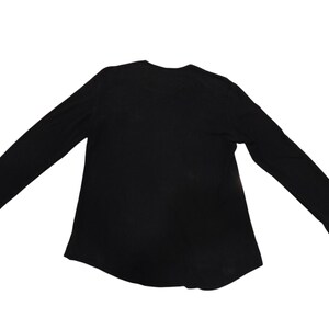 Puede incluir: Una camiseta negra de manga larga con un cuello redondo sencillo y un dobladillo recto. La prenda se presenta sobre un fondo blanco liso, destacando su dise&ntilde;o sencillo, mangas largas y una confecci&oacute;n b&aacute;sica para el uso diario.
