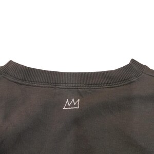 May include: Dark gray sweatshirt with a crew neck and a small, white crown graphic embroidered on the upper back. The sweatshirt has a ribbed neckline and a simple, minimalist design.