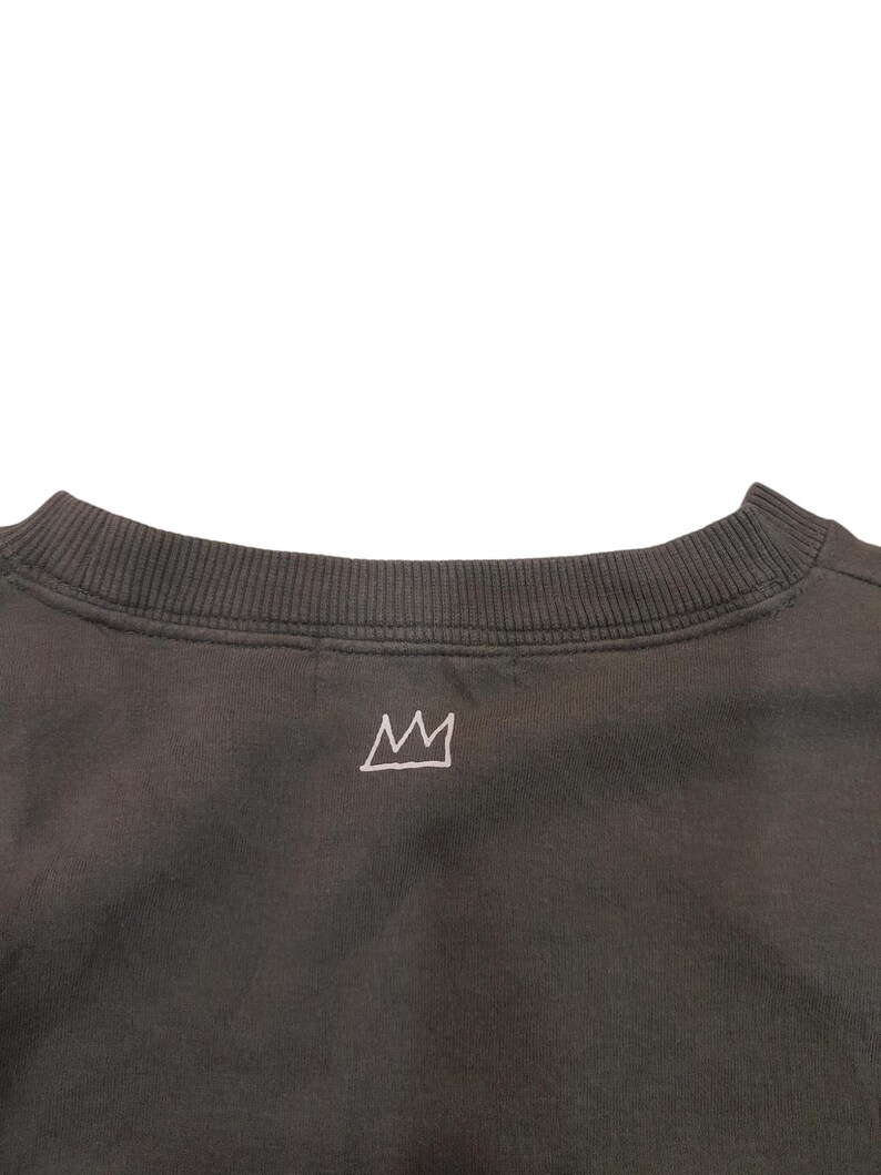 May include: Dark gray sweatshirt with a crew neck and a small, white crown graphic embroidered on the upper back. The sweatshirt has a ribbed neckline and a simple, minimalist design.