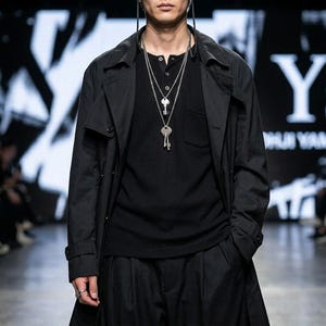 Puede incluir: Una camiseta negra de manga larga tipo henley con bolsillo en el pecho, usada debajo de una gabardina negra. El conjunto se complementa con dos collares plateados superpuestos con colgantes en forma de llave y un anillo de plata.