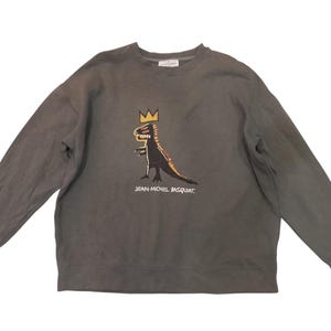 May include: A gray long-sleeved sweatshirt featuring a dinosaur graphic with a crown, in black, yellow, and red. The text "JEAN-MICHEL BASQUIAT" is printed below the graphic. The sweatshirt is made of a soft material.