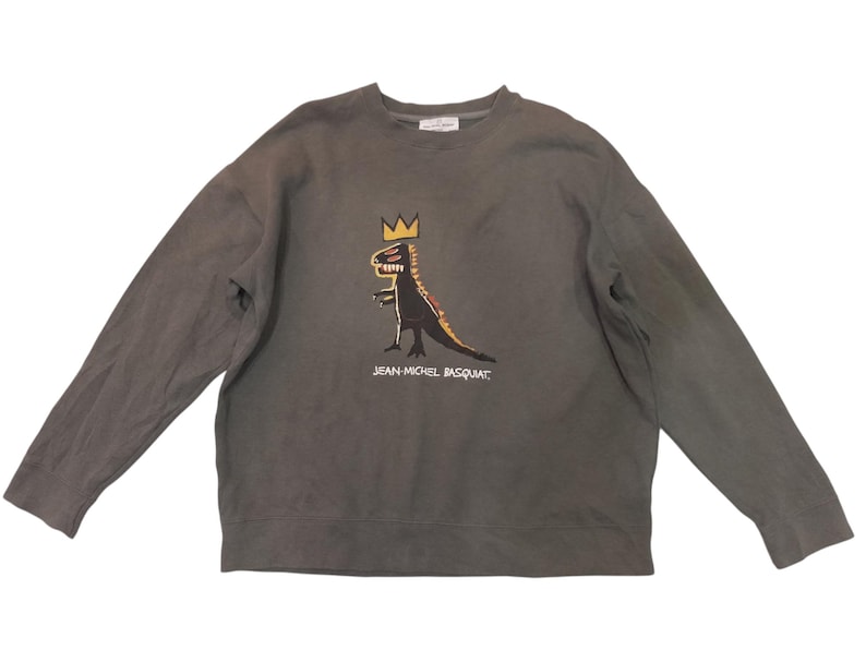 May include: A gray long-sleeved sweatshirt featuring a dinosaur graphic with a crown, in black, yellow, and red. The text "JEAN-MICHEL BASQUIAT" is printed below the graphic. The sweatshirt is made of a soft material.