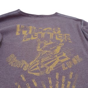 May include: A purple long-sleeve thermal shirt with a distressed yellow graphic print. The design features a skeleton riding a bomb with the text "WINGS LETTER" above and "SKULLS LUCK" below, accompanied by a pattern of small falling bombs.