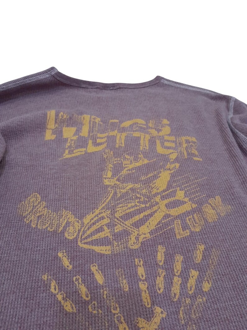 May include: A purple long-sleeve thermal shirt with a distressed yellow graphic print. The design features a skeleton riding a bomb with the text "WINGS LETTER" above and "SKULLS LUCK" below, accompanied by a pattern of small falling bombs.