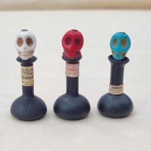 Puede incluir: Tres botellas negras en miniatura con tapones en forma de calavera en blanco, rojo y turquesa. Cada botella presenta una etiqueta de papel con texto impreso, incluyendo las palabras Laudanum, Black Leaf 40 Poison y Poison, con base redonda.