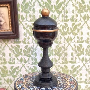 Könnte beinhalten: Eine Miniatur-Dekoleuchte in Schwarz mit einer goldfarbenen Spitze und Zierleiste. Die Lampe steht auf einem kleinen, runden Tisch mit einem farbenfrohen Mosaikdesign. Der Hintergrund zeigt eine grün-weiße Blumentapete.