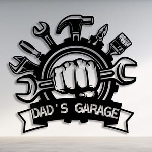 May include: Black metal wall art featuring a fist holding a wrench, surrounded by tools like a hammer, pliers, and screwdriver. The text "DAD'S GARAGE" is displayed on a banner below the tools. The artwork is set against a white wall.
