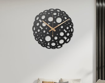 Puede incluir: Un reloj de pared moderno negro y dorado con un diseño circular que presenta un grupo de círculos superpuestos. El reloj está colgado en una pared blanca en un ambiente de sala de estar con un sofá blanco y almohadas marrones.