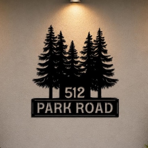 May include: A black metal wall sign featuring a silhouette of five pine trees above a rectangular plaque. The plaque displays the number 512 and the text PARK ROAD in a bold, sans-serif font, mounted centrally on a light-coloured exterior wall.