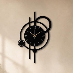 Op de afbeelding: Een moderne zwarte wandklok met een minimalistisch ontwerp. De klok heeft een ronde wijzerplaat met gouden wijzers en een geometrische opstelling van cirkels en lijnen. De klok is bevestigd aan een beige muur.