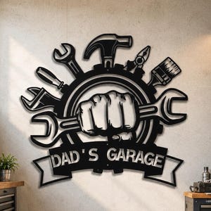 May include: A black metal wall sign featuring a central fist gripping a wrench, surrounded by various tools including a hammer, pliers, and a paintbrush, all arranged within a gear shape. A banner below displays the text DAD'S GARAGE.