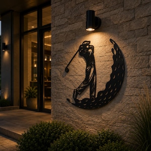 Puede incluir: Una pieza de arte mural de metal negro que presenta la silueta de un golfista en pleno swing, integrada en una forma de media luna llena con un patr&oacute;n circular de hoyuelos que recuerda a una pelota de golf, montada en una pared de piedra.