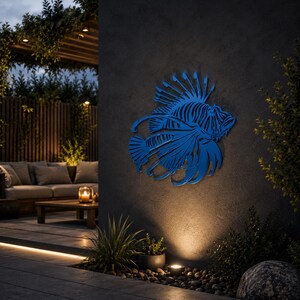 Puede incluir: Una pieza de arte mural de metal azul con forma de pez le&oacute;n con intrincados detalles recortados, que presenta aletas largas y fluidas y una estructura dorsal espinosa, montada sobre una pared exterior gris oscuro con acabado mate.