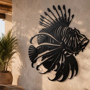 Puede incluir: Una pieza de arte mural de metal negro con forma de pez le&oacute;n, que presenta detalles recortados intrincados que definen las aletas espinosas, la cola fluida y los rasgos faciales distintivos del pez, montada sobre una pared texturizada.