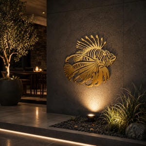 Puede incluir: Una pieza de arte mural de metal dorado con forma de pez le&oacute;n con detalles intrincados y fluidos en las aletas. El dise&ntilde;o recortado presenta espinas afiladas y un cuerpo texturizado, montado sobre una pared de piedra gris oscuro iluminada.