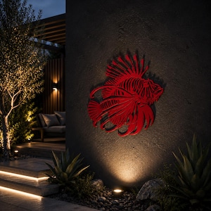 Puede incluir: Una pieza de arte mural de metal rojo vibrante con forma de pez le&oacute;n, que presenta intrincados detalles recortados que definen sus aletas, espinas y estructura corporal. La escultura est&aacute; montada en una pared exterior oscura y texturizada.