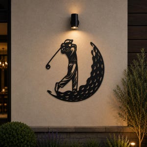 Puede incluir: Una pieza de arte mural de metal negro que presenta la silueta de un golfista en pleno movimiento de swing. El golfista est&aacute; posicionado dentro de una forma de media luna texturizada con hendiduras circulares, imitando una pelota de golf.