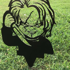 K&ouml;nnte beinhalten: Schwarze Metallsilhouette einer Figur aus dem Horrorfilm Chucky, die ein Messer h&auml;lt. Die Silhouette steht auf einem gr&uuml;nen Rasen.