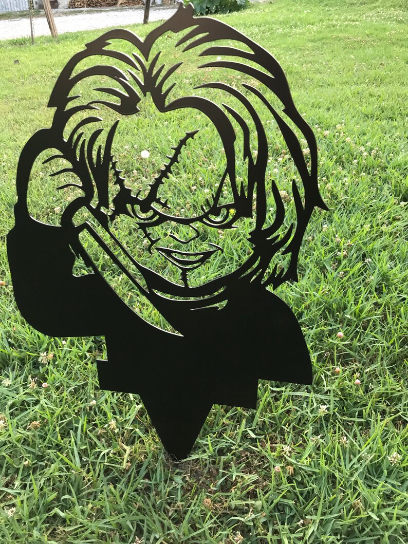K&ouml;nnte beinhalten: Schwarze Metallsilhouette einer Figur aus dem Horrorfilm Chucky, die ein Messer h&auml;lt. Die Silhouette steht auf einem gr&uuml;nen Rasen.