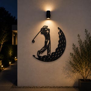 Puede incluir: Una pieza de arte mural de metal negro que presenta la silueta de un golfista en pleno swing, integrada en una forma de media luna con un patr&oacute;n texturizado que recuerda a los hoyuelos de una pelota de golf, montada en una pared exterior clara.