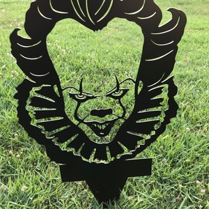 K&ouml;nnte beinhalten: Schwarze Metallsilhouette von Pennywise dem tanzenden Clown aus dem Film ES. Die Silhouette hat die Form eines Kopfes mit einem breiten L&auml;cheln und scharfen Z&auml;hnen. Die Silhouette befindet sich auf einem gr&uuml;nen Grasgrund.