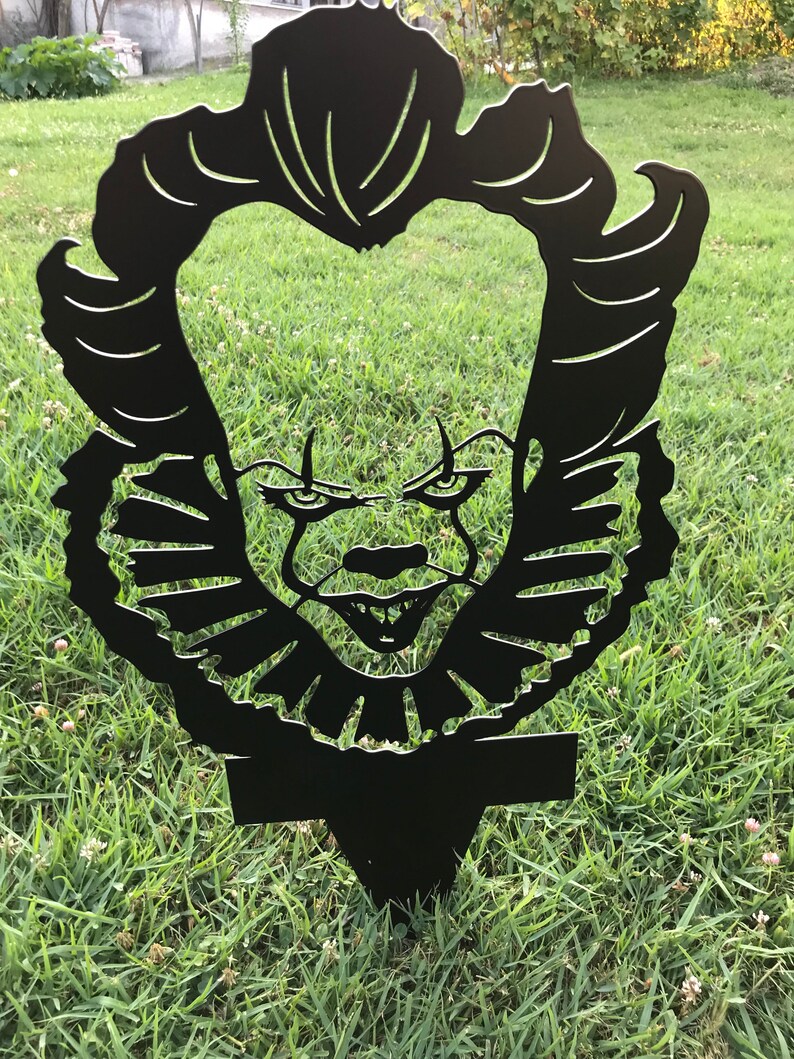 K&ouml;nnte beinhalten: Schwarze Metallsilhouette von Pennywise dem tanzenden Clown aus dem Film ES. Die Silhouette hat die Form eines Kopfes mit einem breiten L&auml;cheln und scharfen Z&auml;hnen. Die Silhouette befindet sich auf einem gr&uuml;nen Grasgrund.