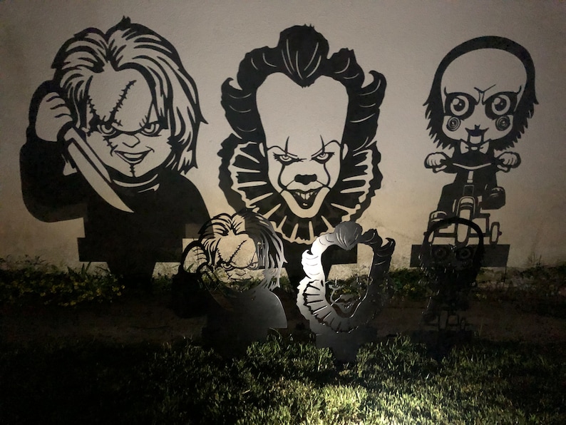 K&ouml;nnte beinhalten: Schwarze Metallausschnitte von Horrorfilmfiguren, darunter Pennywise der tanzende Clown, Chucky und eine Figur aus dem Film Saw. Die Ausschnitte sind an einer Wand montiert und werden von einer Lichtquelle beleuchtet.