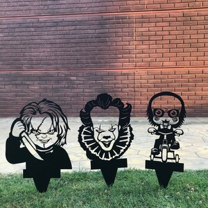 K&ouml;nnte beinhalten: Drei schwarze Metall-Gartenstecker mit den Gesichtern von Horrorfilmfiguren: Pennywise der tanzende Clown, Chucky und eine Figur aus der Saw-Reihe. Die Stecker sind in einem Rasen gesteckt.