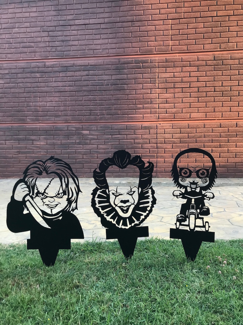 K&ouml;nnte beinhalten: Drei schwarze Metall-Gartenstecker mit den Gesichtern von Horrorfilmfiguren: Pennywise der tanzende Clown, Chucky und eine Figur aus der Saw-Reihe. Die Stecker sind in einem Rasen gesteckt.