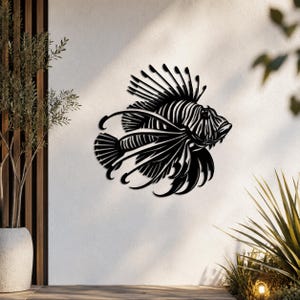 Puede incluir: Una pieza de arte mural de metal negro con forma de pez le&oacute;n, que presenta recortes intrincados que definen las aletas espinosas, el cuerpo rayado y la cabeza detallada del pez. La silueta est&aacute; montada sobre una superficie vertical clara.