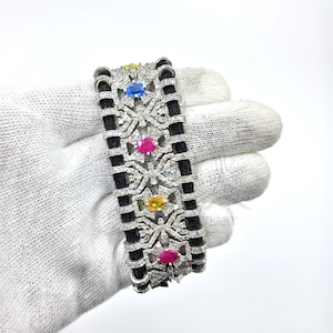 May include: A luxury bracelet featuring a black cord design with intricate silver-tone filigree and sparkling clear stones. The bracelet is accented with vibrant gemstones in yellow, blue, and pink. This elegant piece is perfect for special occasions.