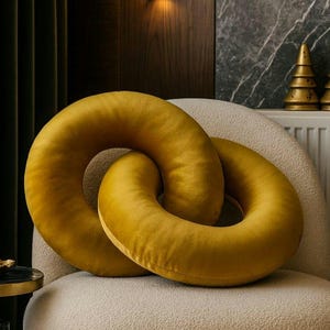 May include: Two mustard-yellow, ring-shaped decorative cushions rest on a cream-coloured, textured chair. The cushions are intertwined, creating a unique design element. The chair has a gold-coloured metal frame. The background features a dark wall and a decorative lamp.
