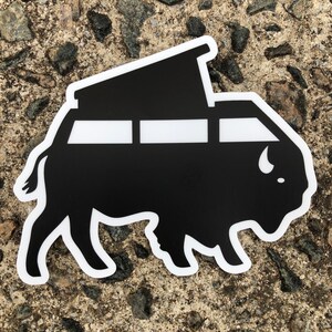 May include: A black and white sticker featuring a stylized bison shape with a camper van design. The van has three rectangular windows and a pop-up roof. The sticker is on a textured, gray surface.