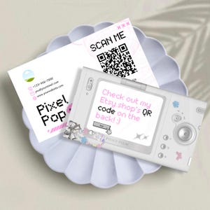 Puede incluir: Una tarjeta de visita blanca con el texto "Pixel Pop" y la información de contacto descansa sobre un plato festoneado. Una cámara digital blanca con el texto "Check out my Etsy shop's QR code on the back! :)" está junto a una tarjeta con un código QR.
